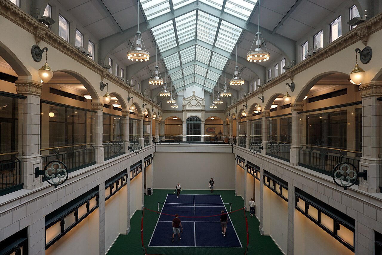 Players on a pickleball court with white lines on a blue surface, set up on the floor of a skylit historic arcade atrium